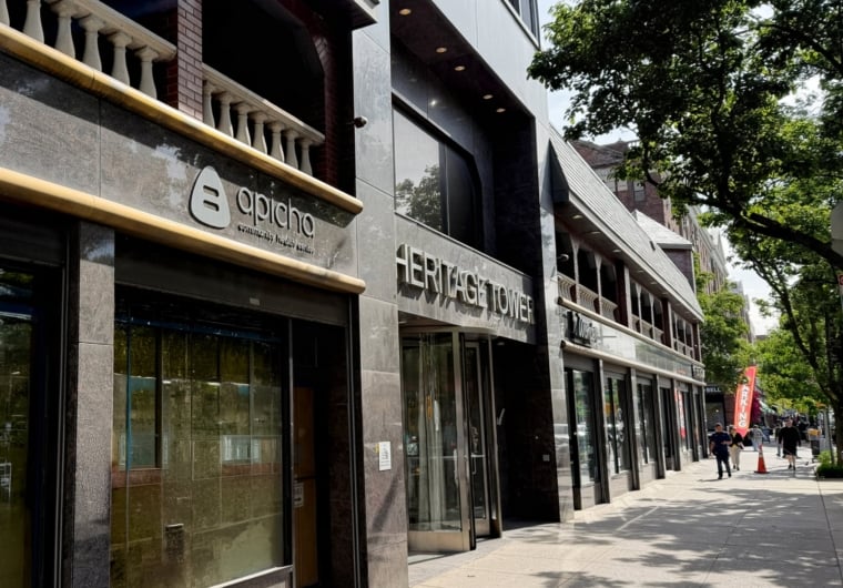 Street view of Heritage Tower building with glass entrance and adjacent apicho restaurant on a sunny day. Trees line the sidewalk, and a few people walk in the distance.