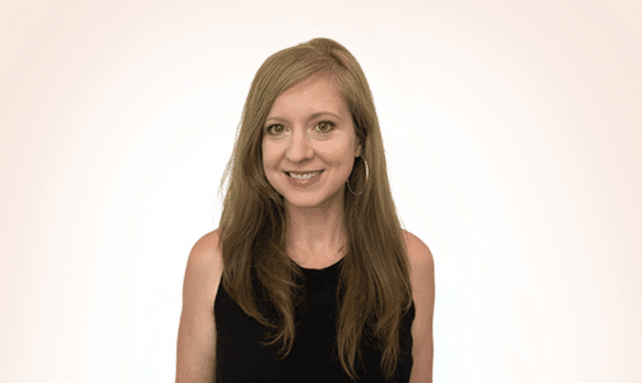 A woman with long light brown hair, wearing a sleeveless black top and hoop earrings, stands in front of a plain light background, smiling at the camera.