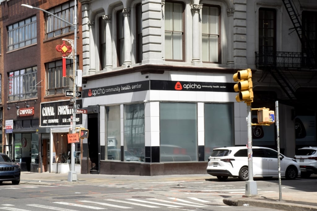 Corner view of Apicha Community Health Center with its sign visible, located at a city intersection with traffic lights, parked cars, and surrounding storefronts.