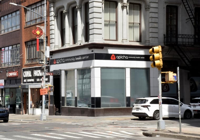 Corner view of Apicha Community Health Center with its sign visible, located at a city intersection with traffic lights, parked cars, and surrounding storefronts.