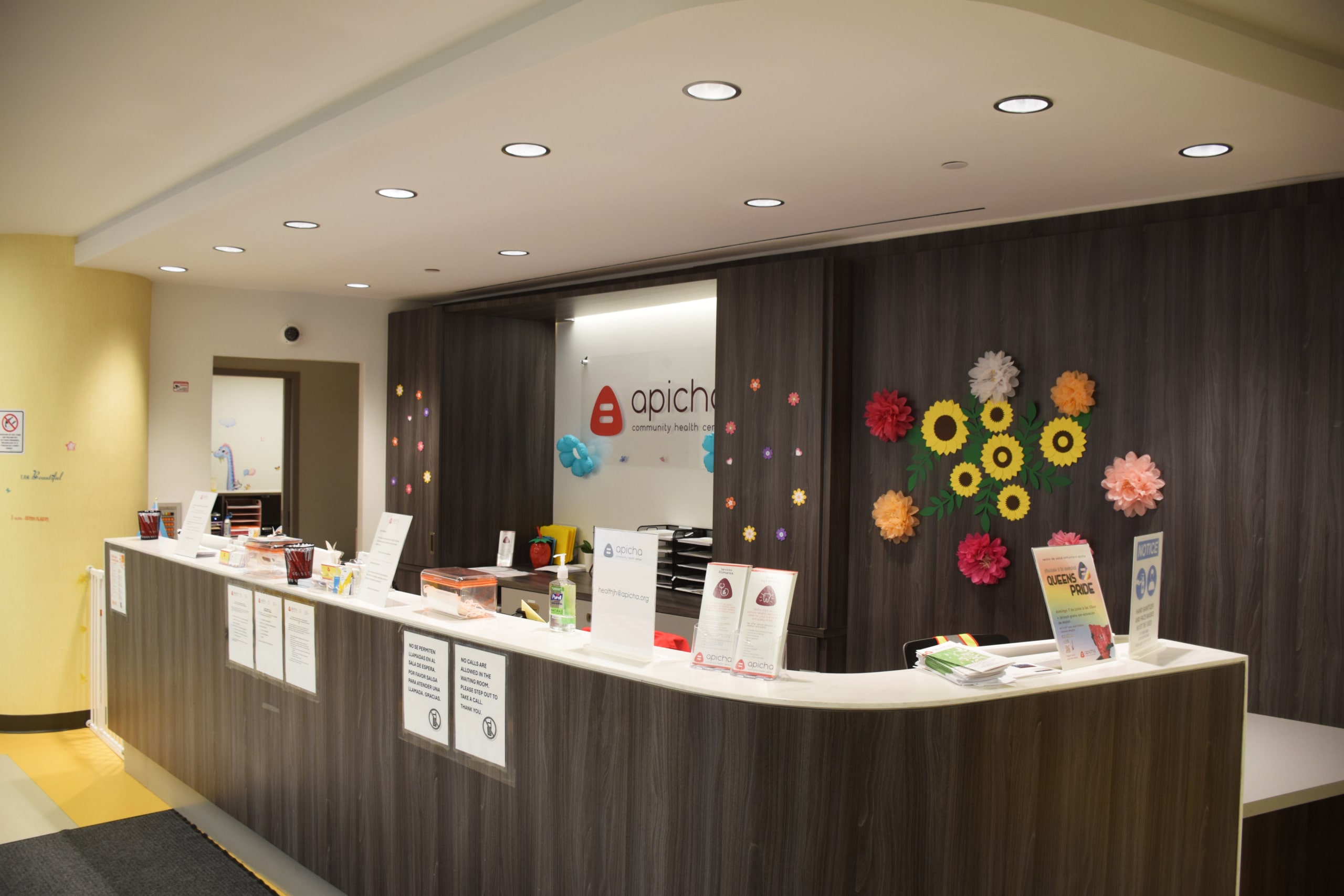 A modern reception desk with informational signs, colorful paper flowers on the wall, and the APICHA Community Health Center logo behind the counter.