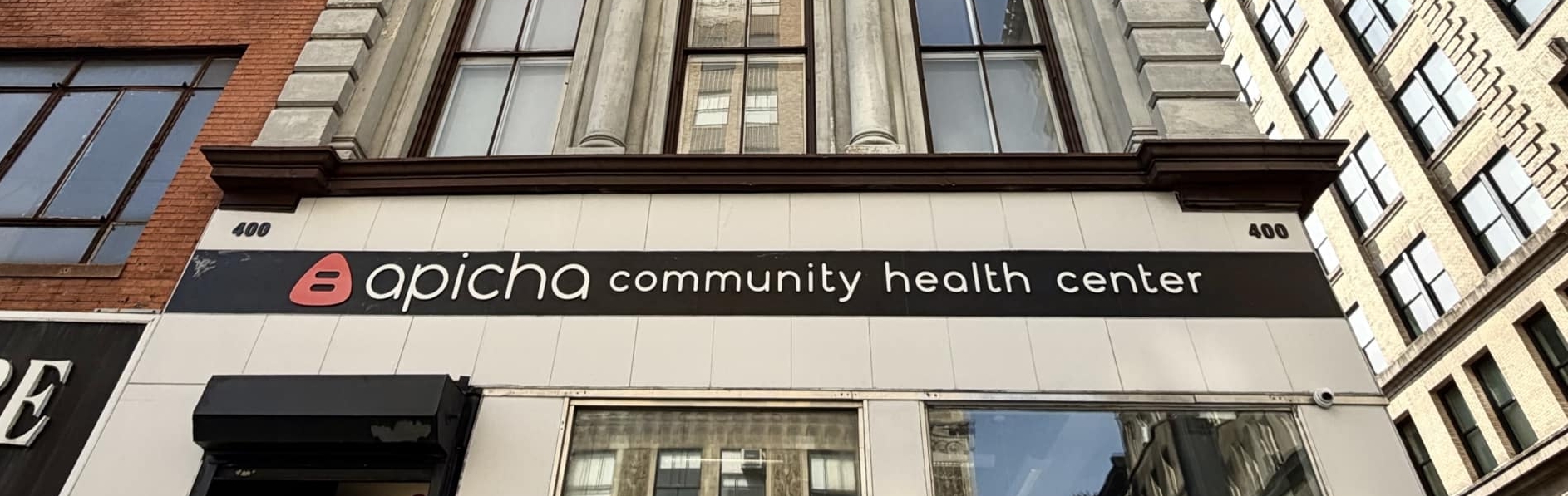 A multi-story building houses Apicha Community Health Center, with large windows and a sign visible above the entrance on a city street.
