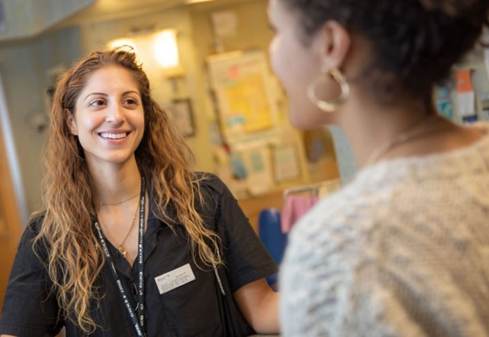 A woman wearing scrubs and a name badge smiles while speaking to another woman in a casual sweater inside a brightly lit indoor setting.