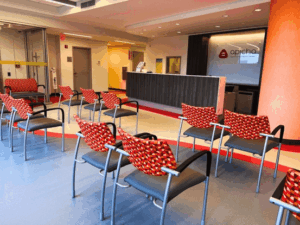Empty clinic waiting room with patterned chairs facing a reception desk and a sign that reads "apicha health center" on the back wall.