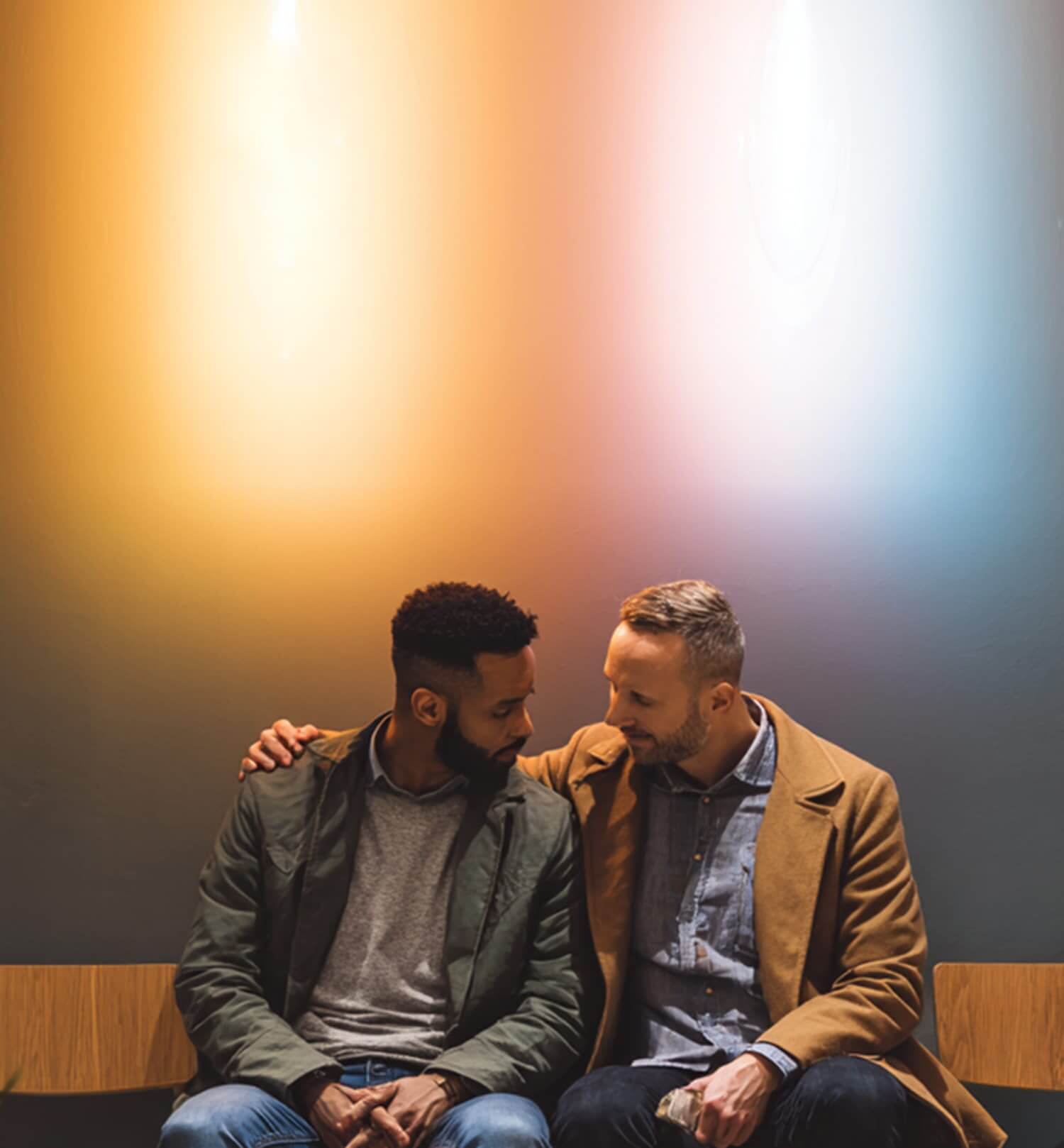 Two men sit side by side on a bench, one with his arm around the other. Warm, orange and blue lighting illuminates the wall behind them.