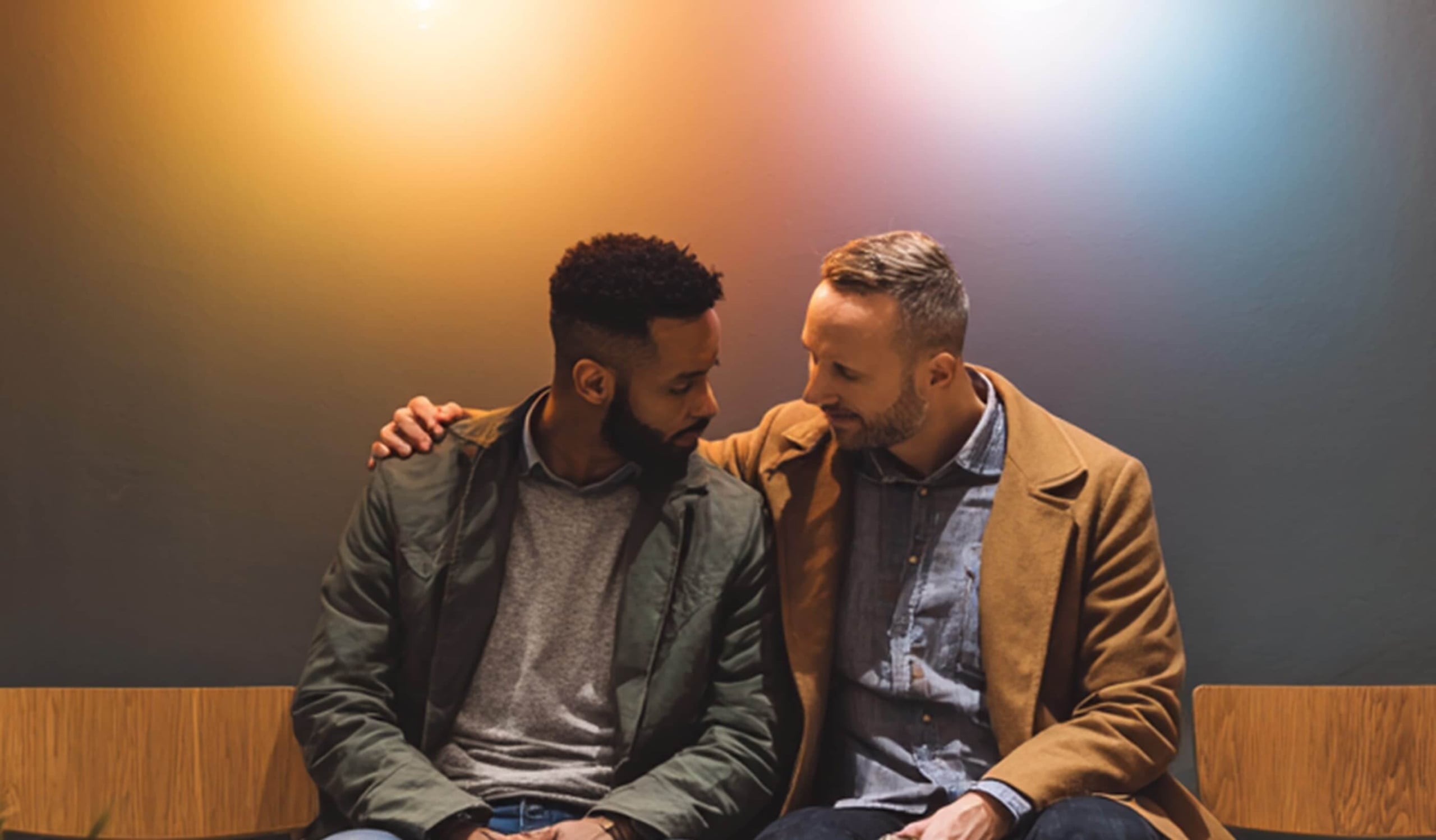 Two men sitting closely on a bench, one with his arm around the other, both looking at each other under warm, soft lights against a plain background.