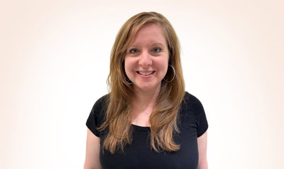 Woman with long light brown hair wearing a black short-sleeve top, smiling, standing against a plain white background.