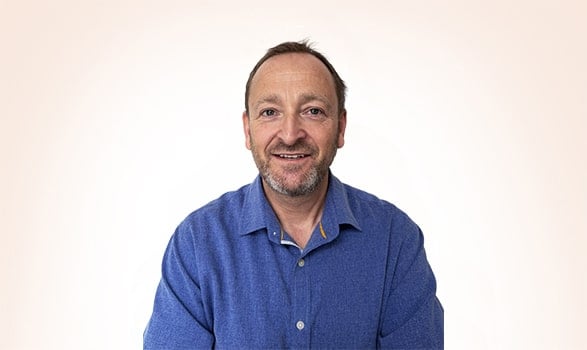 Man with short brown hair and beard wearing a blue button-up shirt, smiling, posed against a plain light background.
