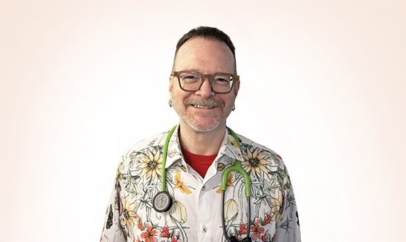 A smiling person wearing glasses, a floral shirt, and a stethoscope around their neck, standing against a plain light background.