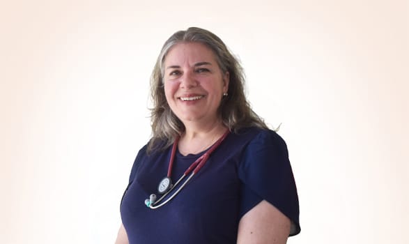 A woman with gray hair wearing a navy blue top and a stethoscope around her neck stands smiling against a plain light background.