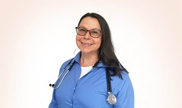 Woman wearing glasses and a blue shirt with a stethoscope around her neck, smiling at the camera against a plain light background.