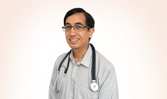 A man wearing glasses and a light gray shirt with a stethoscope around his neck stands against a plain white background.