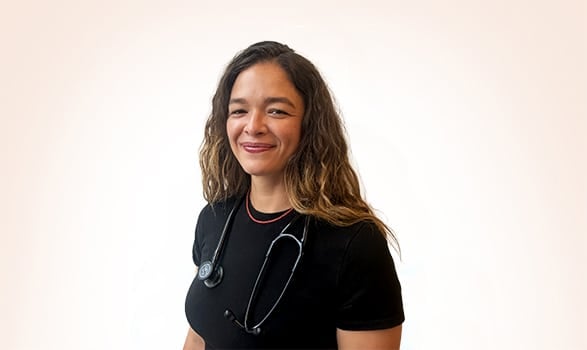 A woman with long wavy hair wearing a black shirt and a stethoscope around her neck stands against a plain light background, smiling at the camera.