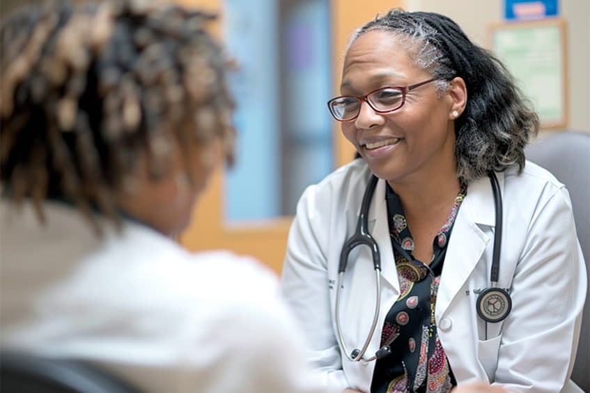 A doctor wearing a white coat and stethoscope sits and talks with a patient in a medical office.