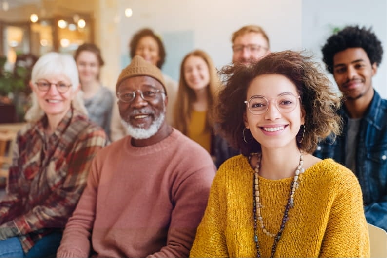 A diverse group of people sitting closely together indoors, smiling and looking at the camera.