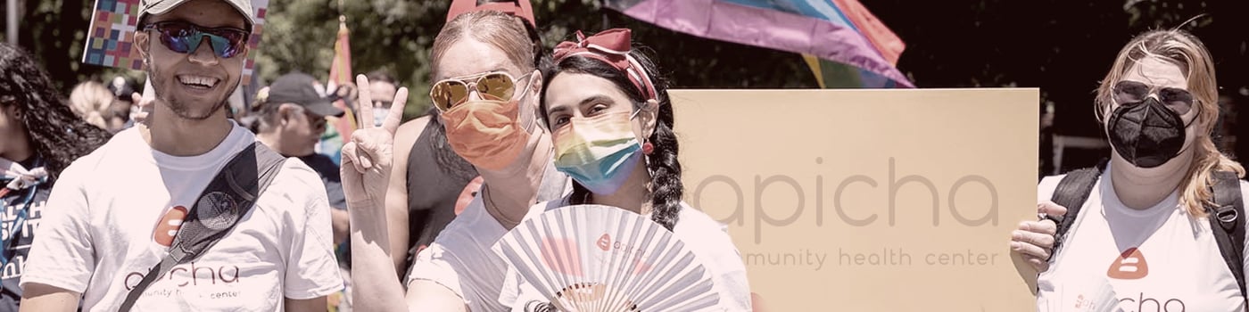 Four people wearing Apicha t-shirts and face masks stand together at an outdoor event, holding a fan and a large yellow sign, with rainbow flags visible in the background.