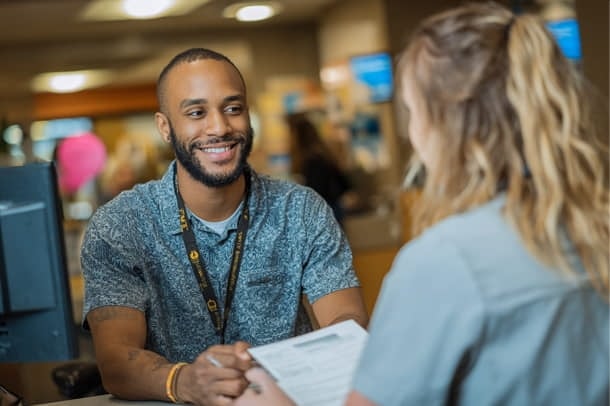A man at a service desk smiles while talking to a woman holding paperwork. They appear to be in a professional or customer service setting.