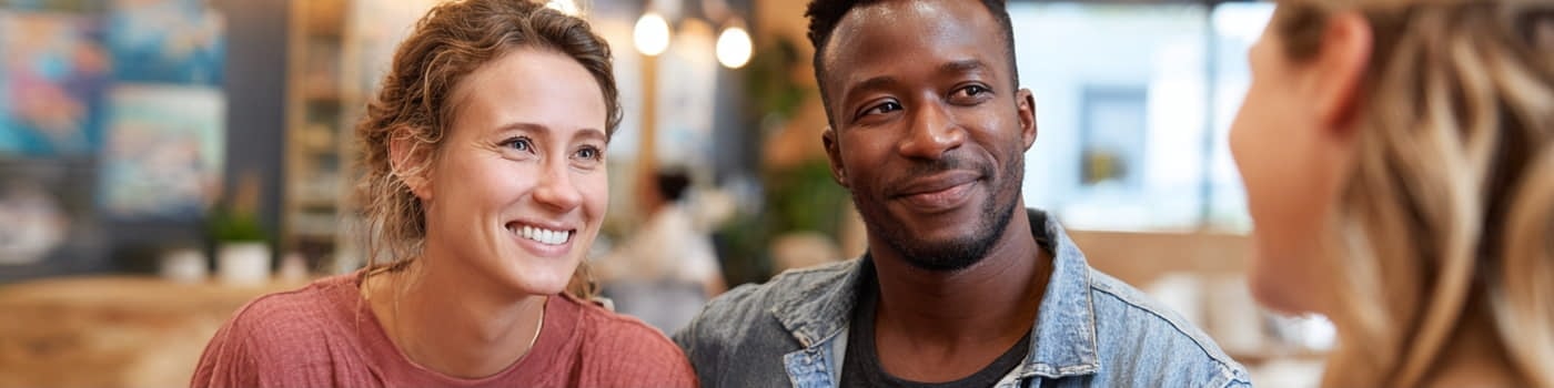 Two people sit close together and smile while talking with another person in a well-lit indoor setting.