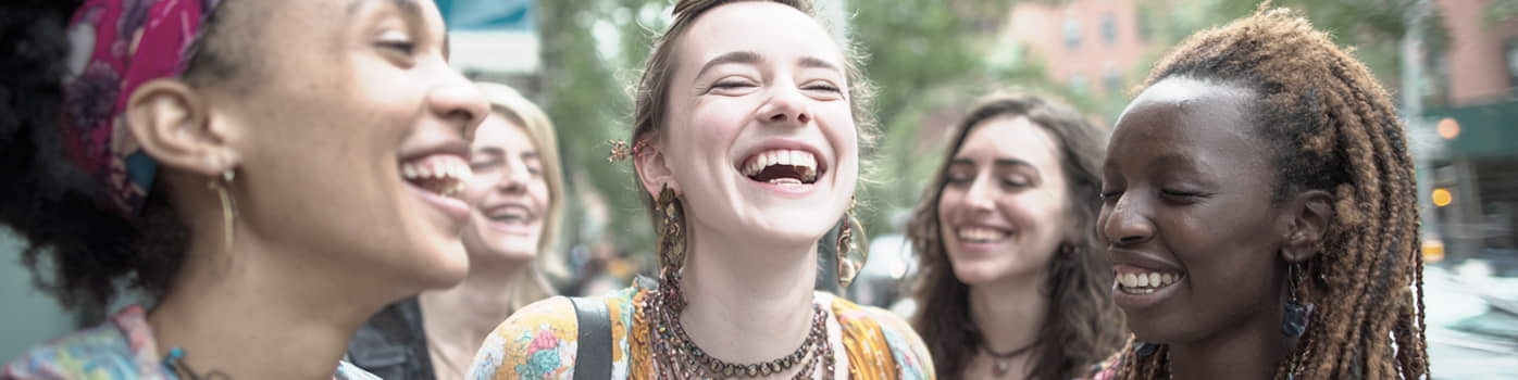 Five young women standing outdoors, smiling and laughing together on a city street.