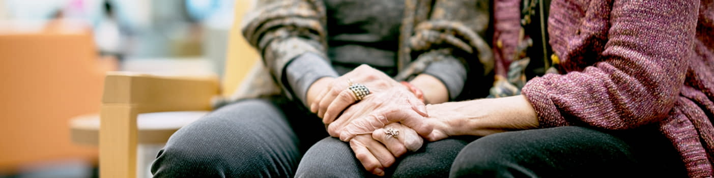 Two older adults sit side by side on a bench, holding hands for support; their faces are not visible, and only their torsos and hands are shown.