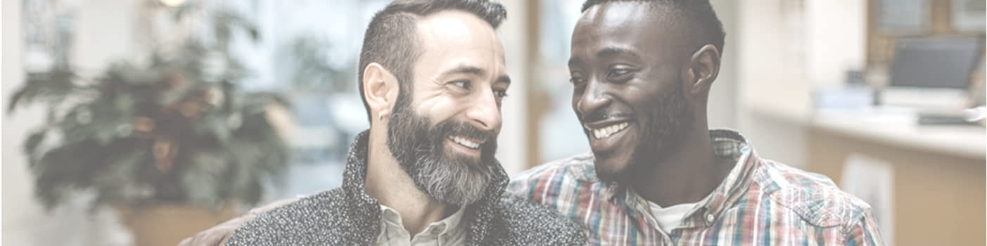 Two men sit close together indoors, smiling and looking at each other.
