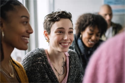 A group of people sit together indoors, smiling and talking. The focus is on a person with short hair wearing a sweater and necklace.