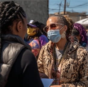 Two women talk outdoors at a public event; one wears sunglasses, a mask, and a patterned jacket, while others stand in the background.