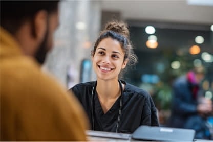 A woman in black scrubs sits at a desk, smiling and talking to a person in the foreground. A laptop is on the desk and the background appears to be an indoor public space.