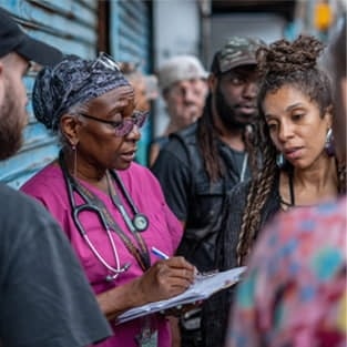 A healthcare worker in scrubs and a stethoscope writes on a clipboard while talking to a group of people outdoors.