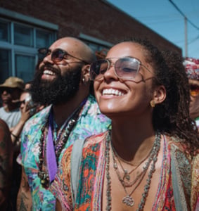 Two people wearing sunglasses and layered necklaces smile while standing outdoors at a daytime event, with other people and a brick building in the background.