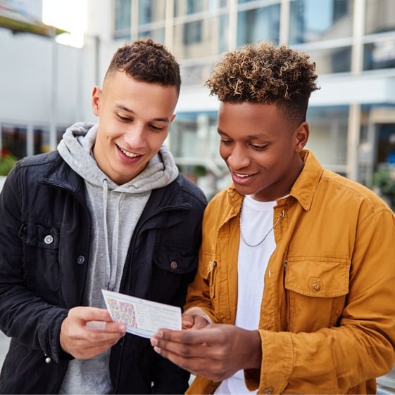 Two young men stand outside, smiling as they look at a colorful printed paper together.