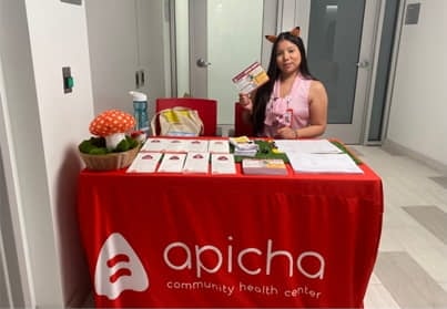 A woman sits at an information table covered with a red "apicha community health center" tablecloth, holding flyers. Various pamphlets and a mushroom decoration are on the table.