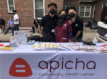 Three people wearing masks stand behind an Apicha Community Health Center table with brochures and supplies at an outdoor event.