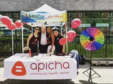 Four people stand under a tent at an outdoor event booth for Apicha Community Health Center, with balloons, a prize wheel, and informational materials on the table.