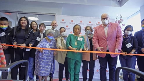 A group of people, some wearing masks, stand behind a ribbon at a ribbon-cutting ceremony indoors.