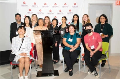 A group of people pose for a photo indoors, standing and sitting in front of a branded backdrop with red logos. A podium is positioned at the front center.