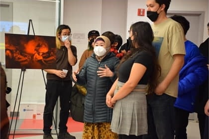 A group of people wearing masks stand indoors near an easel with artwork; one person in a puffer jacket and hat speaks while others listen attentively.