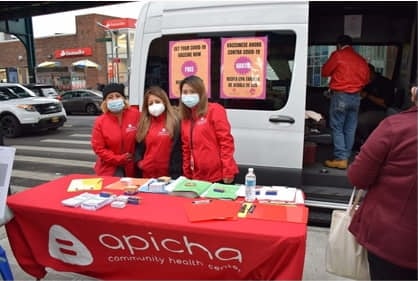 Three people in red jackets and masks stand behind a table with health materials and an "apicha community health center" banner, next to a van on a city street.