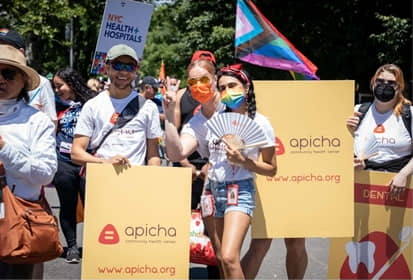 A group of people hold yellow Apicha signs and wear masks at an outdoor event, with a Pride flag and a NYC Health + Hospitals sign visible in the background.