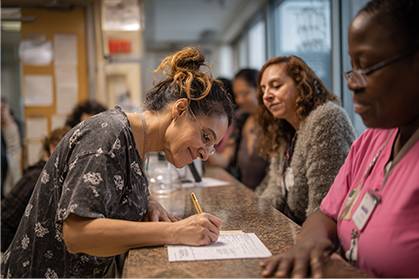 A woman fills out paperwork at a reception desk while two women, including a staff member in pink scrubs, observe.