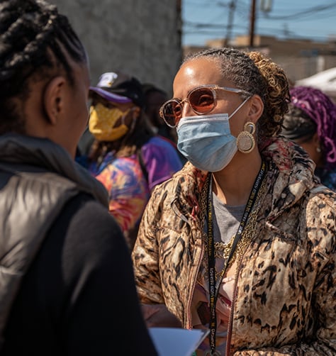 Two women wearing face masks talk outdoors in a crowd. One wears sunglasses and a patterned jacket; people and power lines are visible in the background.