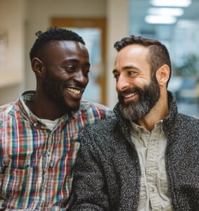 Two men sitting close together indoors, smiling at each other. One wears a plaid shirt, the other has a beard and wears a textured jacket.