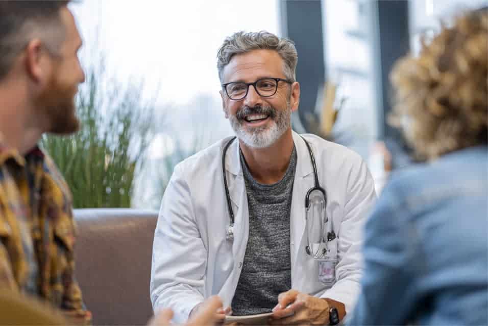 A smiling doctor with a stethoscope around his neck talks to two patients in a bright, modern office.