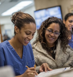 Two women sit at a desk, one in blue scrubs writing on a form while the other, in glasses and a light sweater, smiles and looks on.
