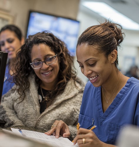 Two women, one in blue scrubs and the other in a sweater, sit together at a desk smiling while reviewing and signing paperwork.