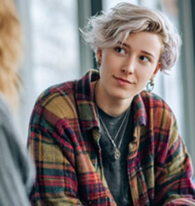 A person with short, wavy light hair wearing a plaid shirt and layered necklaces looks attentively at someone off camera.