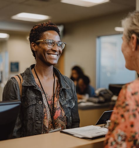 A woman wearing glasses and a denim jacket smiles while speaking to another person at a reception desk in an office setting.