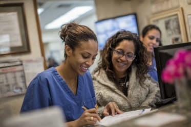 Two women talk at a medical reception desk while one in blue scrubs fills out paperwork and another person stands behind a computer monitor.
