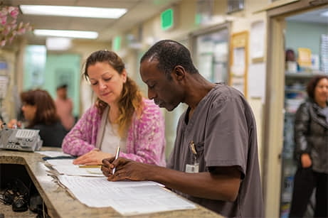 A man in scrubs and a woman review documents together at a hospital reception desk, with other people and staff in the background.