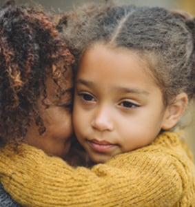 An adult and a child with curly hair share a close hug. The adult has their face against the child's cheek. The child looks calm and thoughtful, wearing a yellow sweater.